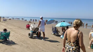 Un vendedor ambulante vestido completamente de blanco empuja un carro con una gran nevera por la arena de la playa de la Victoria de Cádiz, rodeado de bañistas y sombrillas junto al mar.