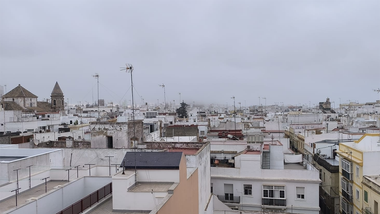 Vista panorámica de Cádiz desde una azotea del barrio de La Viña en una mañana cubierta por la niebla. En primer plano se ven las azoteas y edificios del casco antiguo, mientras la espesa bruma reduce notablemente la visibilidad y oculta en la distancia algunos de los principales elementos del perfil de la ciudad, como la Catedral y el segundo puente.