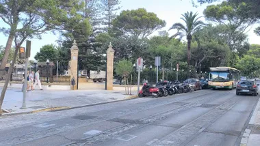 Entrada al Parque Genovés de Cádiz durante una mañana soleada y despejada, con varias personas caminando, motocicletas y coches estacionados y un autobús circulando por la calzada.
