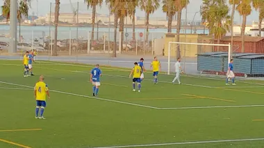 Imagen panorámica del partido entre los equipos de veteranos del Cádiz CF y del San Fernando. Varios jugadores con camisetas amarillas y azules se encuentran dentro del área mientras disputan una acción ofensiva. Al fondo se observan palmeras, la Bahía de Cádiz y las instalaciones portuarias, en una estampa característica del entorno del complejo deportivo Puntales-La Paz.