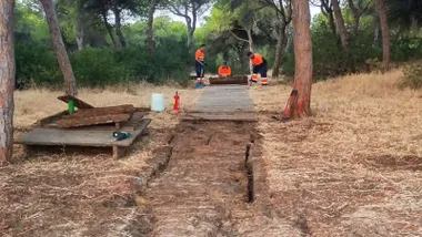 Pasarela de madera siendo reparada por unos operarios en mitad del acceso boscoso a la playa.