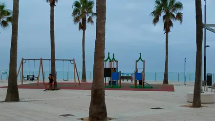 Plaza de la Estrella de Cádiz, junto a la playa de Santa María del Mar, con palmeras, un parque infantil casi vacío y el cielo completamente cubierto.