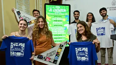 Grupo de siete personas posa durante la presentación de ‘El Encuentro’ en Cádiz. Varias sostienen camisetas azules y blancas del Día de la Juventud y banderines con el lema «¿Podemos hablar?». En el centro aparece una pantalla vertical con el cartel y la programación de ‘El Encuentro Andalucía’ del 29 de agosto en la Plaza de España de Cádiz.
