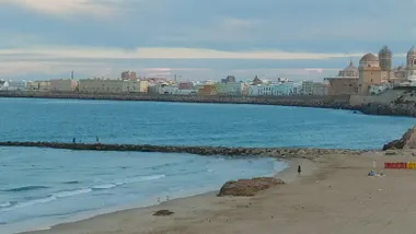 Vista panorámica tomada desde la playa de Santa María del Mar hacia el casco histórico de Cádiz en la mañana del lunes 3 de agosto. En la imagen se distinguen la Catedral y el Campo del Sur bajo un cielo cubierto de nubes, con los últimos restos de calima antes de su desaparición. El mar está en calma y la playa presenta poca afluencia de personas, reflejando el ambiente más fresco tras el descenso de las temperaturas registrado en la provincia.