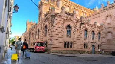Dos personas arrastran una maleta amarilla y otra azul por una calle de Cádiz, con la trasera rosada del Gran Teatro Falla a su derecha y un cielo azul despejado sobre los edificios.