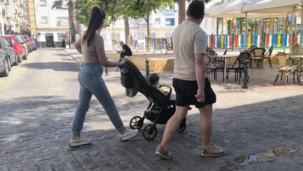 Una pareja camina con un coche de bebé por la plaza Manolo Santander de Cádiz, con una zona de juegos infantiles y terrazas al fondo.