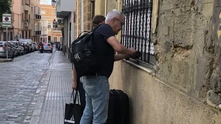 Dos turistas con equipaje, uno de ellos con una gran maleta, sacan una llave de una caja de seguridad con código instalada junto a la ventana de un edificio en una calle de Cádiz.