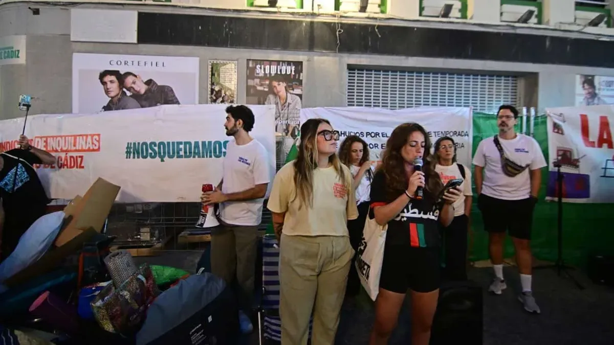 Varias personas participan en la asamblea de El Palillero, una de ellas hablando con un micrófono, junto a pancartas del Sindicato de Inquilinas y enseres utilizados durante la acampada.