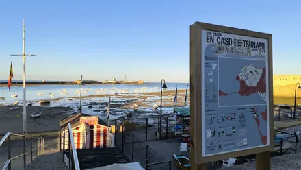 Cartel de recomendaciones en caso de tsunami en la playa de la Caleta de Cádiz, con el Castillo de San sebastián al fondo y bajo un cielo azul y despejado