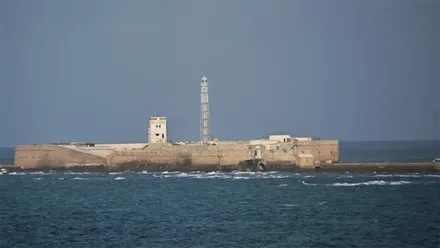 Vista del Castillo de San Sebastián desde la costa de Cádiz, rodeado por el mar, con las murallas de la fortaleza y el faro elevándose en el centro del recinto.