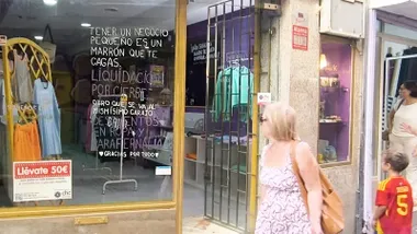 Varias personas pasan junto al escaparate de La Chula, en los Callejones de Cardoso de Cádiz, donde grandes mensajes manuscritos sobre el cristal anuncian la liquidación por cierre de la tienda.