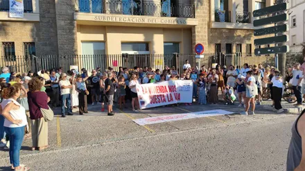 Decenas de personas concentradas frente a la Subdelegación del Gobierno en Cádiz sostienen una gran pancarta con el lema «La vivienda nos cuesta la vía», mientras una portavoz se dirige a los asistentes con un megáfono.