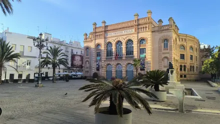 Vista del Gran Teatro Falla de Cádiz desde la plaza Fragela, con su característica fachada de ladrillo, palmeras en primer plano y un cielo azul completamente despejado.