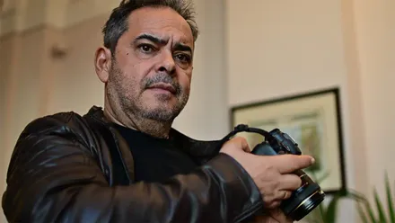 Retrato de Julio González sosteniendo una cámara fotográfica entre las manos. Viste una chaqueta oscura y mira directamente hacia la cámara. Al fondo, desenfocados, se distinguen un cuadro y parte de una estancia interior.