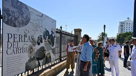 El alcalde de Cádiz, Bruno García, y la presidenta del puerto, Teófila Martínez, junto a varias personas observan un gran panel expositivo instalado junto a la verja del muelle, en Cádiz. En primer plano aparece el cartel "Perlas del Caribe" y, al fondo, se distinguen palmeras y edificios de la ciudad.