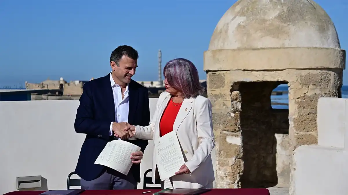 Bruno García y Blanca Flores se estrechan la mano mientras sostienen los documentos del acuerdo, con elementos de la fortificación y el mar al fondo.