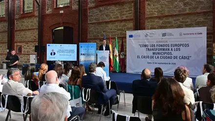 Asistentes sentados ante un escenario en el interior de los antiguos Depósitos de Tabaco de Cádiz, donde un ponente interviene durante la jornada 'Cómo los fondos europeos transforman a los municipios. El caso de la ciudad de Cádiz'.