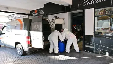 Dos operarios con epi blanco cargando un bidón azul con comida en mal estado.