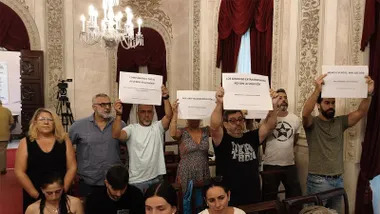 Varias personas participan en una protesta dentro del salón de plenos del Ayuntamiento de Cádiz. Algunas sostienen carteles con mensajes como “Cumplimiento total Acuerdo Regulador”, “Por una valoración justa”, “Los servicios extraordinarios no son la solución” y “Menos eventos, más gestión”.