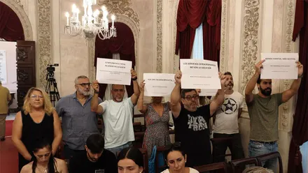 Varias personas participan en una protesta dentro del salón de plenos del Ayuntamiento de Cádiz. Algunas sostienen carteles con mensajes como “Cumplimiento total Acuerdo Regulador”, “Por una valoración justa”, “Los servicios extraordinarios no son la solución” y “Menos eventos, más gestión”.
