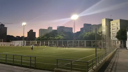 Vista al anochecer de un campo de fútbol de césped artificial con jugadores y dos grandes torretas de iluminación encendidas. Al fondo, muy próximos al terreno de juego, se observan varios bloques de viviendas iluminados por los focos.