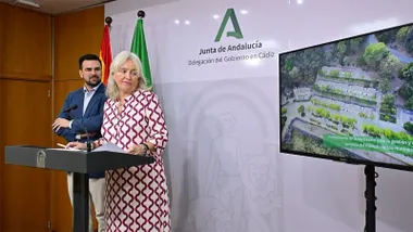 Mercedes Colombo interviene ante un atril junto a Francisco Moreno en la Delegación del Gobierno de la Junta en Cádiz. A su derecha, una pantalla muestra una vista aérea del Poblado de Los Hurones.
