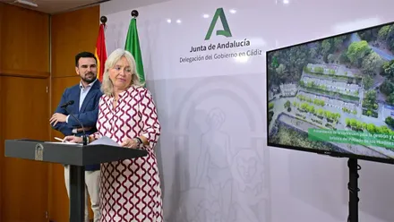 Mercedes Colombo interviene ante un atril junto a Francisco Moreno en la Delegación del Gobierno de la Junta en Cádiz. A su derecha, una pantalla muestra una vista aérea del Poblado de Los Hurones.