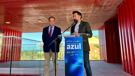 Dos hombres participan en la presentación de Blue Zone Forum InnovAzul 2026. Uno interviene ante un micrófono desde un atril azul con el lema «Proyectando en azul», mientras el otro permanece a su lado, en un espacio de estructura roja y abierto al exterior.