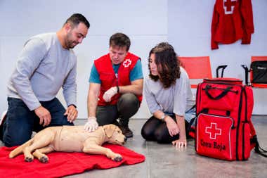 Tres personas participan en una práctica de primeros auxilios con un maniquí de perro tumbado sobre una manta roja. En el centro, un instructor con chaleco de Cruz Roja realiza una maniobra sobre el animal mientras los participantes observan. A la derecha aparece una mochila roja de emergencias.
