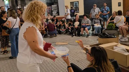Una mujer sirve un trozo de tortilla a otra durante la cena colectiva en la Plaza del Palillero, mientras numerosos asistentes permanecen reunidos al fondo.