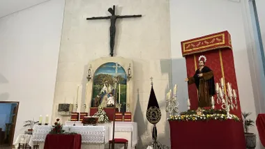 Imagen de San Francisco de Asís situada en un altar con dos candelabros y un exorno floral, ante un dosel rojo, en el interior de un templo. Al fondo se observa el altar mayor con una imagen mariana y un crucificado.