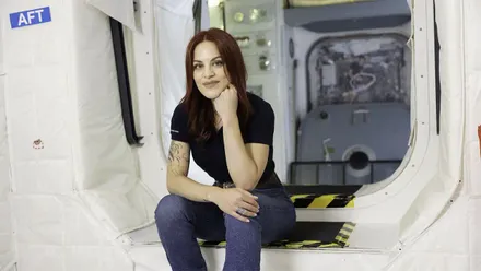 Sara García Alonso aparece sentada en el interior de unas instalaciones de entrenamiento espacial, mirando a cámara y apoyando el rostro sobre una mano. Viste camiseta oscura y pantalón gris, y al fondo se observa una estructura que simula el interior de un módulo espacial.