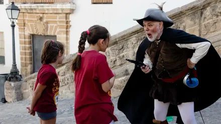 Dos niñas con camisetas de La Carrera del Orbe escuchan a un actor caracterizado con vestuario de época durante una prueba de la yincana en la plaza de Santa Cruz, junto a la Catedral Vieja de Cádiz.