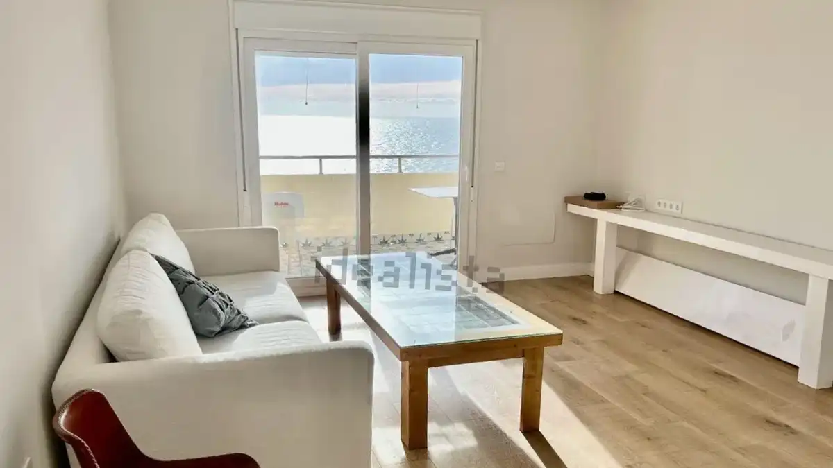 Salón luminoso con sofá blanco y mesa de centro situado frente a un ventanal desde el que se ve directamente el mar.