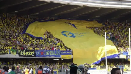 Grada del estadio de Chapín repleta de aficionados del Cádiz CF durante el partido del ascenso, con una gigantesca camiseta del Cádiz CF amarilla y azul desplegada sobre el público y pancartas de peñas cadistas.