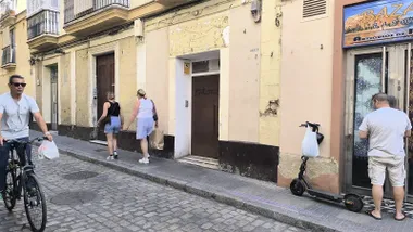 Fachada de un edificio del centro de Cádiz con varias puertas a pie de calle y la marca de un antiguo rótulo comercial sobre una de ellas. Varias personas caminan por la acera y un hombre pasa en bicicleta.