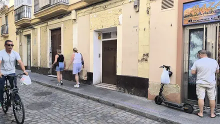 Fachada de un edificio del centro de Cádiz con varias puertas a pie de calle y la marca de un antiguo rótulo comercial sobre una de ellas. Varias personas caminan por la acera y un hombre pasa en bicicleta.