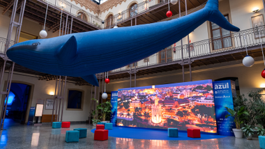 Una enorme figura de ballena azul cuelga del techo de un patio interior rodeado de galerías con barandillas metálicas. Bajo ella hay una pantalla de gran formato con una imagen nocturna de la Torre del Oro de Sevilla, varios asientos cúbicos de colores y carteles del Blue Zone Forum InnovAzul.