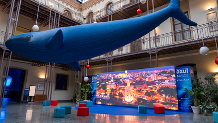 Una enorme figura de ballena azul cuelga del techo de un patio interior rodeado de galerías con barandillas metálicas. Bajo ella hay una pantalla de gran formato con una imagen nocturna de la Torre del Oro de Sevilla, varios asientos cúbicos de colores y carteles del Blue Zone Forum InnovAzul.