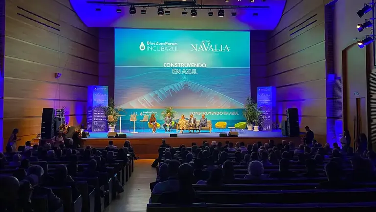 Vista general del auditorio del Palacio de Congresos de Cádiz, con numerosas personas sentadas frente a un escenario iluminado en tonos azules. Sobre el escenario intervienen varios participantes y una gran pantalla muestra los logotipos de Blue Zone Forum y Navalia junto al lema 'Construyendo en azul'.
