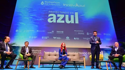 Cinco participantes en el escenario del Blue Zone Forum InnovAzul 2026. Sara García Alonso, vestida con un traje azul, permanece sentada en el centro, mientras un hombre interviene de pie y otros tres escuchan sentados. Detrás, una gran pantalla muestra el lema "Proyectando en azul".