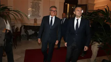 Manuel Gavira y Bruno García, vestidos con trajes oscuros y corbatas, caminan por el interior del Ayuntamiento de Cádiz, sobre una alfombra roja y entre plantas decorativas.