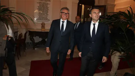 Manuel Gavira y Bruno García, vestidos con trajes oscuros y corbatas, caminan por el interior del Ayuntamiento de Cádiz, sobre una alfombra roja y entre plantas decorativas.