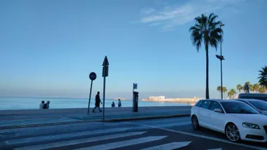 Vista de un paseo marítimo de Cádiz en una mañana despejada, con varias personas caminando junto al mar, palmeras, un paso de peatones en primer plano y un coche blanco a la derecha.