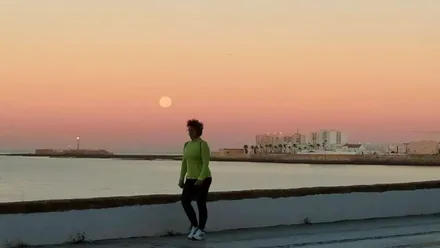 Luna casi llena sobre el mar al amanecer, con una persona caminando por el paseo marítimo de Sancti Petri y la ciudad de Cádiz al fondo.