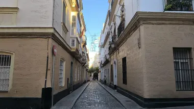 Calle estrecha y empedrada del centro histórico de Cádiz, flanqueada por edificios de fachadas claras con balcones, ventanas enrejadas y faroles. Al fondo se aprecia una zona iluminada por el sol bajo un cielo azul, mientras varias personas caminan por la calle.