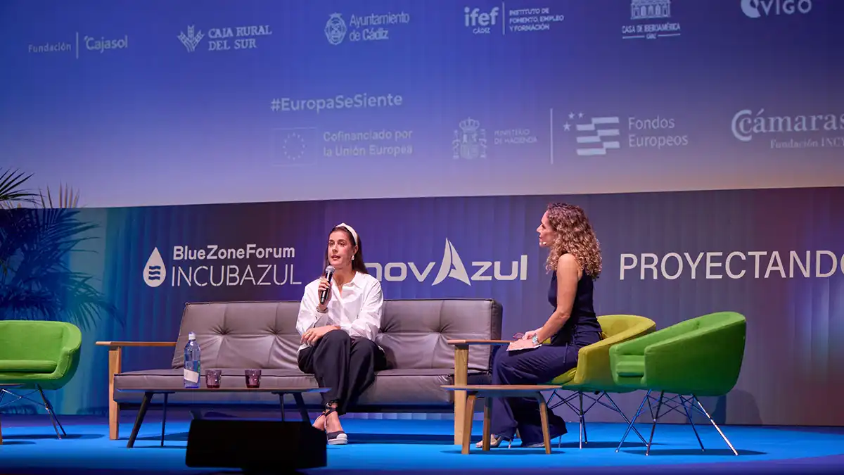 Carolina Marín, vestida con una camisa blanca y pantalón oscuro, habla por un micrófono sentada en un sofá gris. Frente a ella, una entrevistadora de cabello rizado la escucha desde un sillón verde. Ambas están sobre el escenario del Blue Zone Forum InnovAzul 2026, con los logotipos del encuentro al fondo.