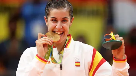 Carolina Marín sonríe mientras muerde su medalla de oro olímpica y sostiene en la otra mano el recuerdo de los Juegos de Río 2016. Viste una chaqueta blanca de la selección española con detalles rojos y amarillos, y lleva la medalla colgada del cuello.