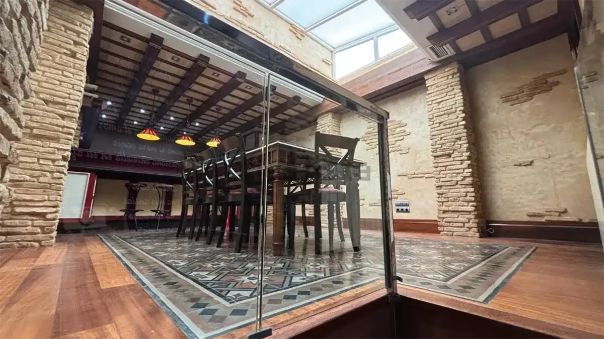 Estancia interior con paredes de ladrillo visto, suelo decorado, cerramiento de cristal, mesa con sillas y entrada de luz natural desde la parte superior.