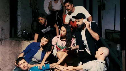 Varios intérpretes aparecen agrupados alrededor de un hombre tumbado en el suelo, en una escena teatral ambientada en un espacio industrial con iluminación tenue.